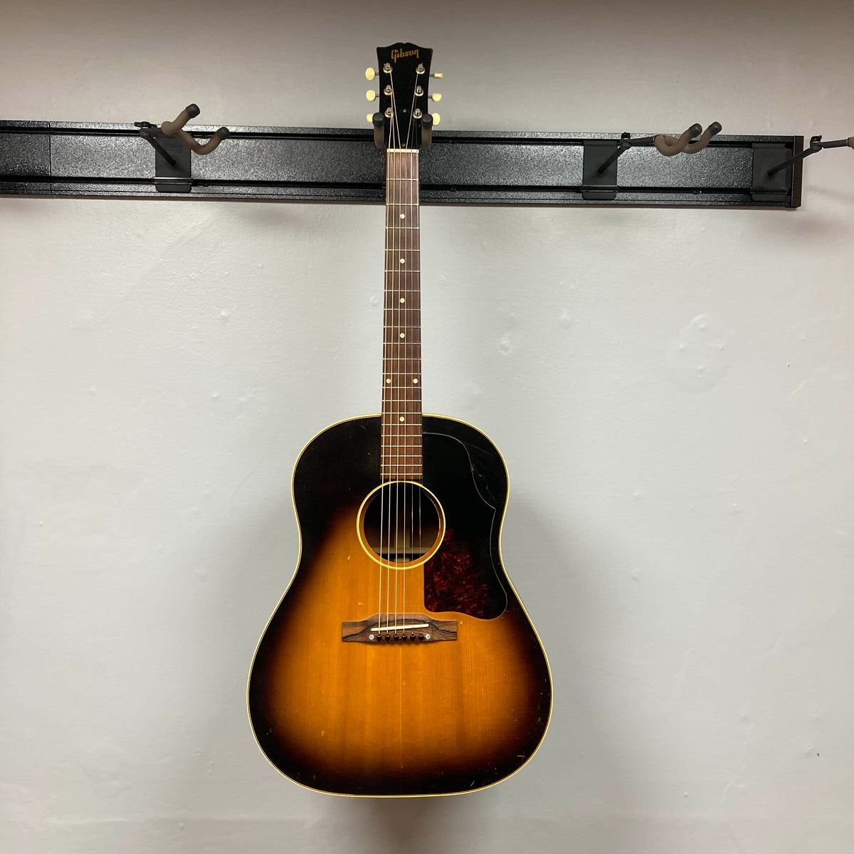 A Gibson J-45 1957 Serial U-8664-1 acoustic guitar by Gibson with a sunburst finish and pickguard hangs on a wall-mounted rack against a plain white background.