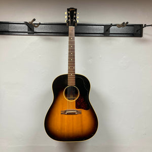 A Gibson J-45 1957 Serial U-8664-1 acoustic guitar by Gibson with a sunburst finish and pickguard hangs on a wall-mounted rack against a plain white background.