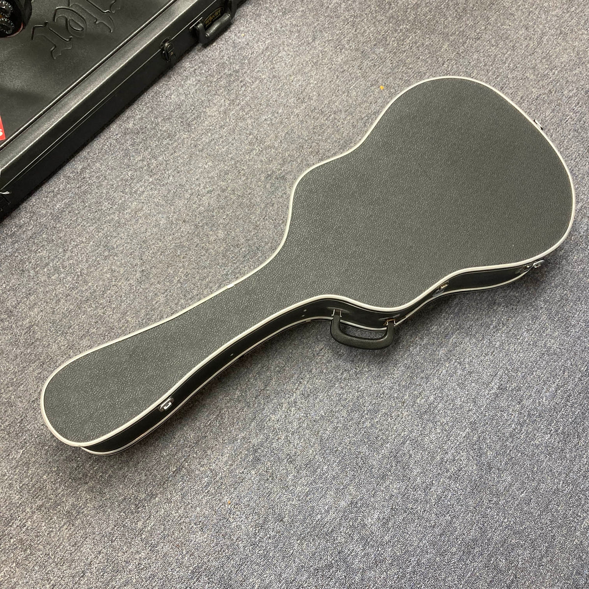 A closed Gibson J-45 1957 (Serial U-8664-1) vintage black guitar case with white trim rests on a gray carpeted floor.
