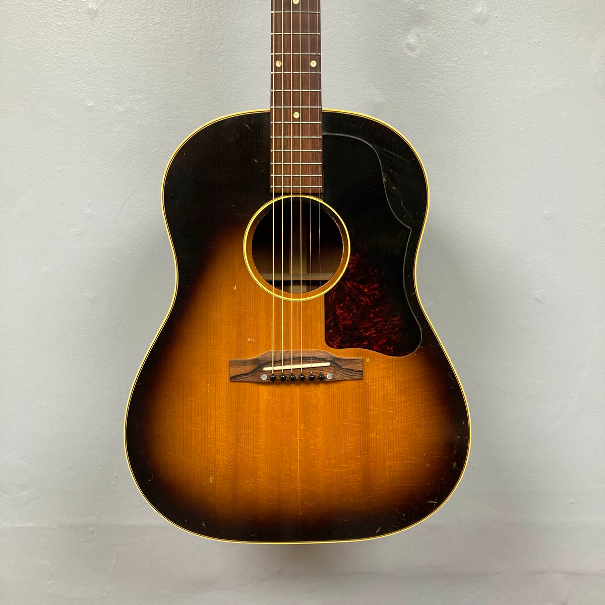 The Gibson J-45 1957 Serial U-8664-1, a vintage acoustic guitar with a sunburst finish, slope shoulder design, visible pickguard, and six strings, is photographed against a plain light gray wall.