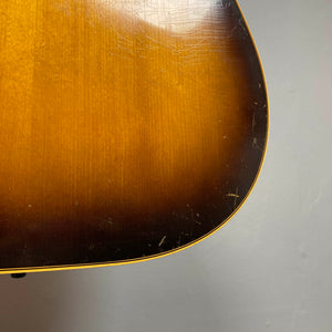 Close-up of the lower body of a Gibson J-45 1957 Serial U-8664-1 acoustic guitar, featuring a sunburst finish, visible surface scratches, and a light-colored outline set against a plain background.