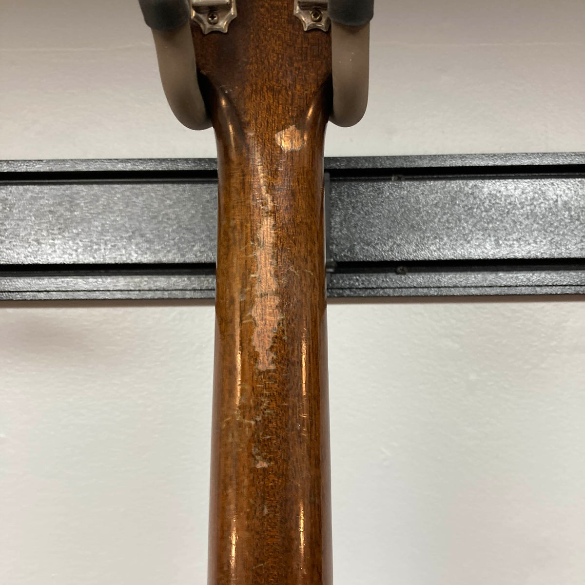 Close-up of the back of a Gibson J-45 1957 Serial U-8664-1 acoustic guitar neck by Gibson, showing visible wear and scratches, mounted on a black wall bracket against a white background.