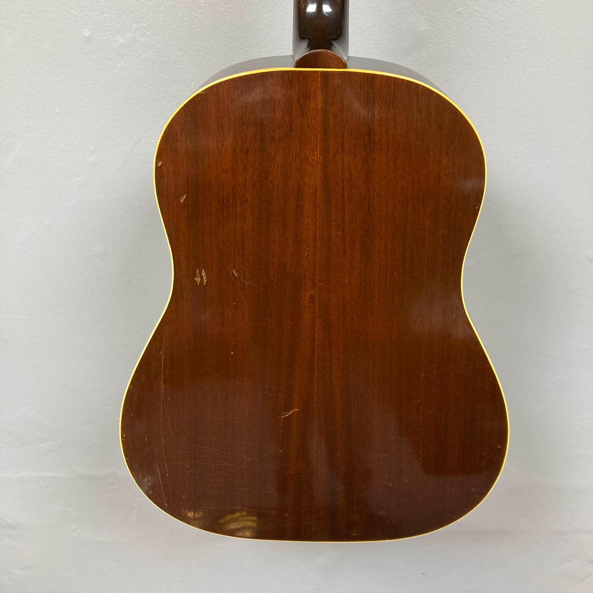 Back view of a Gibson J-45 1957 (Serial U-8664-1) acoustic guitar with a glossy finish, visible scratches, and yellow edge binding. The classic slope shoulder design is set against a light-colored wall.