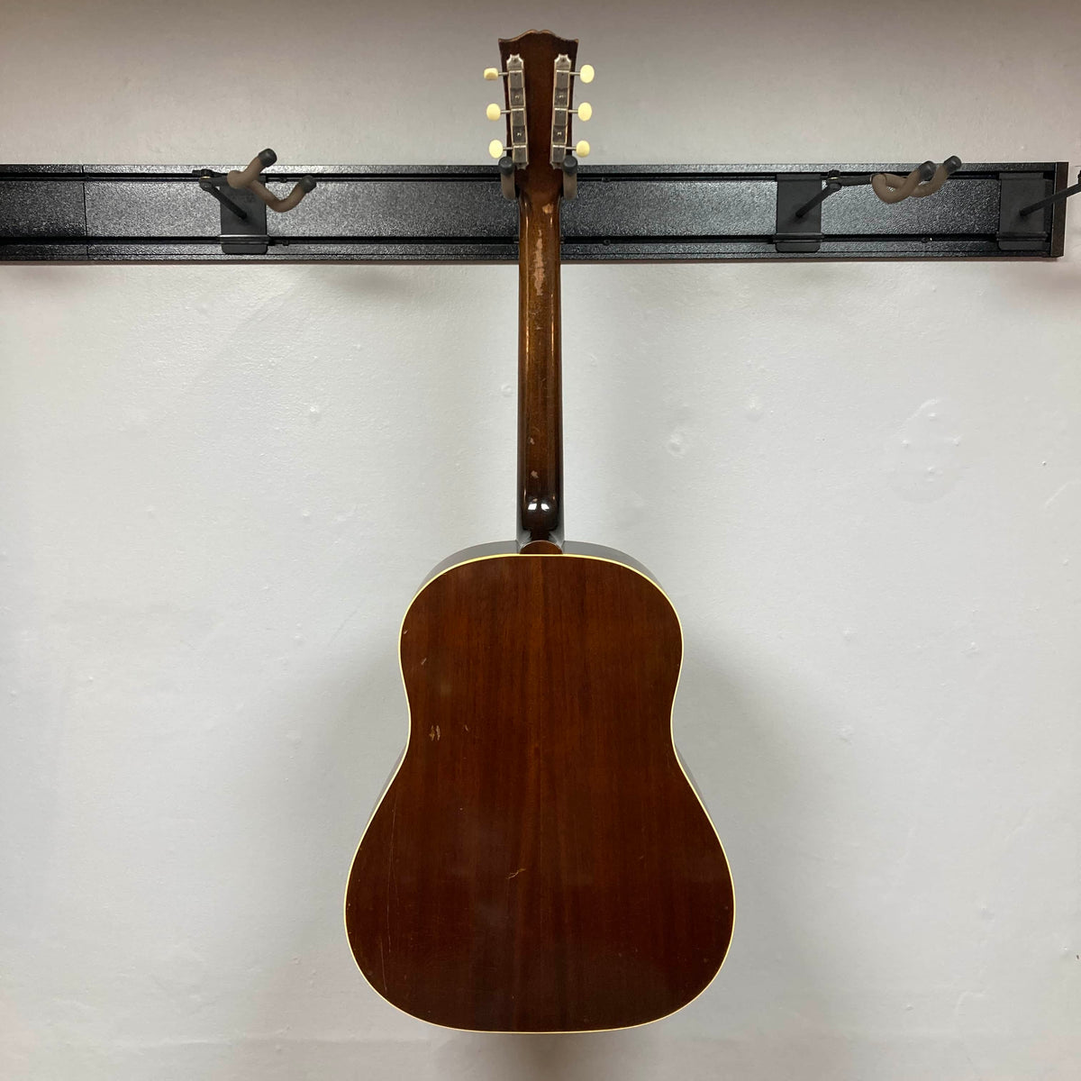 A Gibson J-45 1957 (Serial U-8664-1) vintage acoustic guitar with a slope shoulder design hangs on a wall-mounted rack, shown from the back with the Gibson tuning pegs visible at the top.