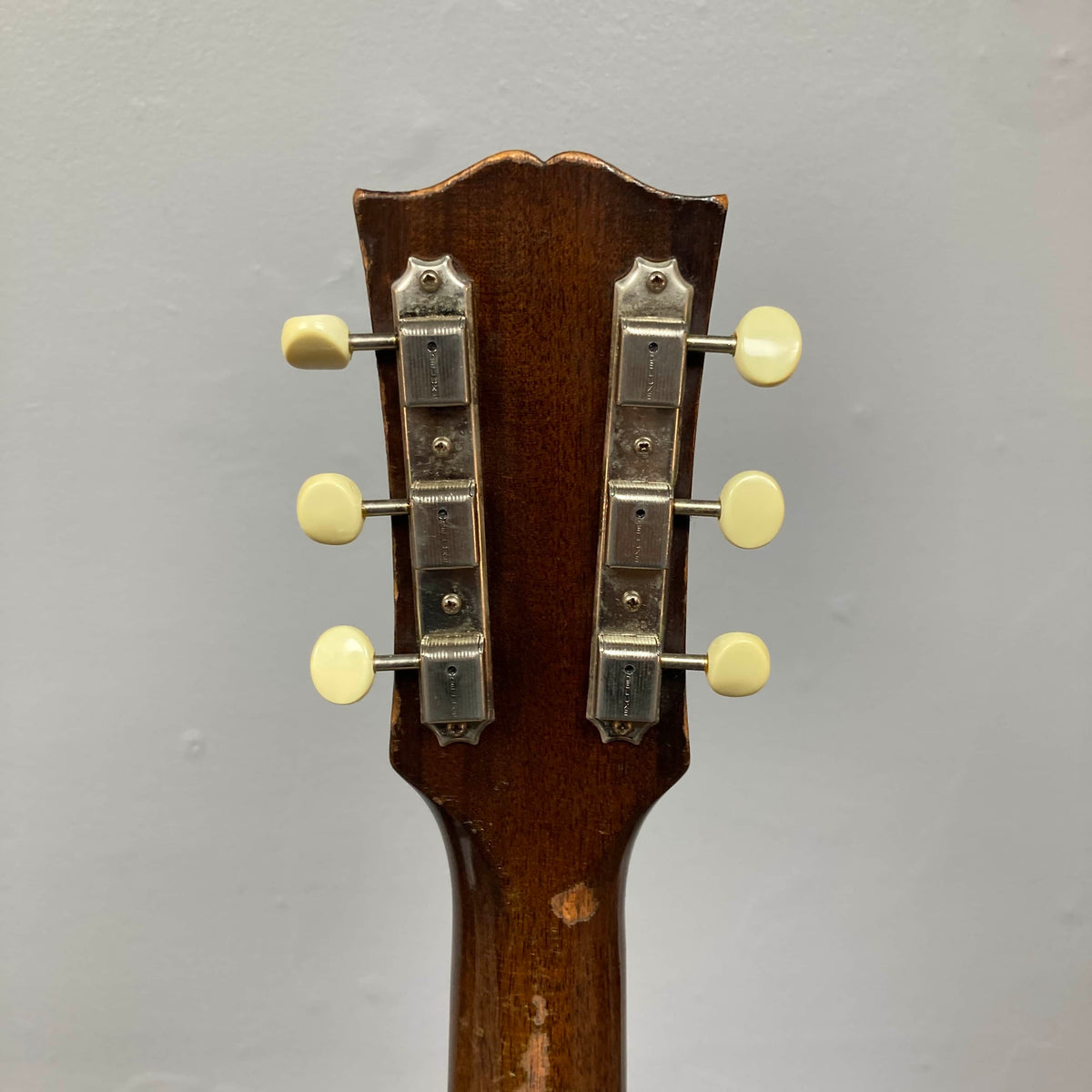 Close-up of the back of a Gibson J-45 1957 (Serial U-8664-1) headstock, showing six cream-knobbed tuning pegs, set against a plain gray background.