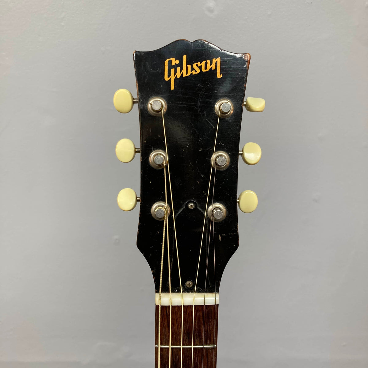 Close-up of the Gibson J-45 1957 Serial U-8664-1 headstock, featuring six tuning pegs with cream buttons and the Gibson logo, shown on a black background to highlight its classic slope shoulder design.