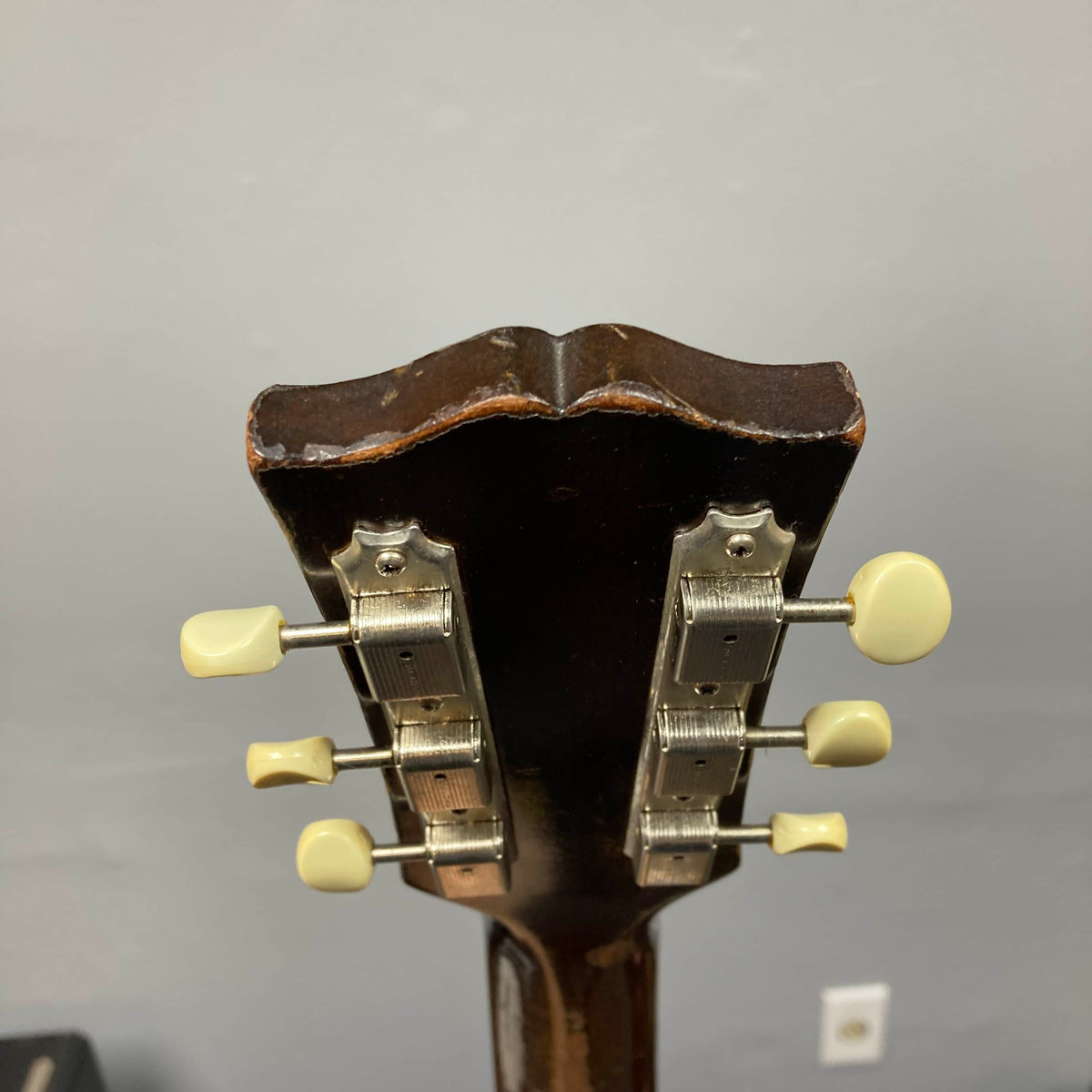 The back of a Gibson J-45 1957 (Serial U-8664-1) headstock, featuring six tuning pegs—three per side—is displayed against a plain gray wall.