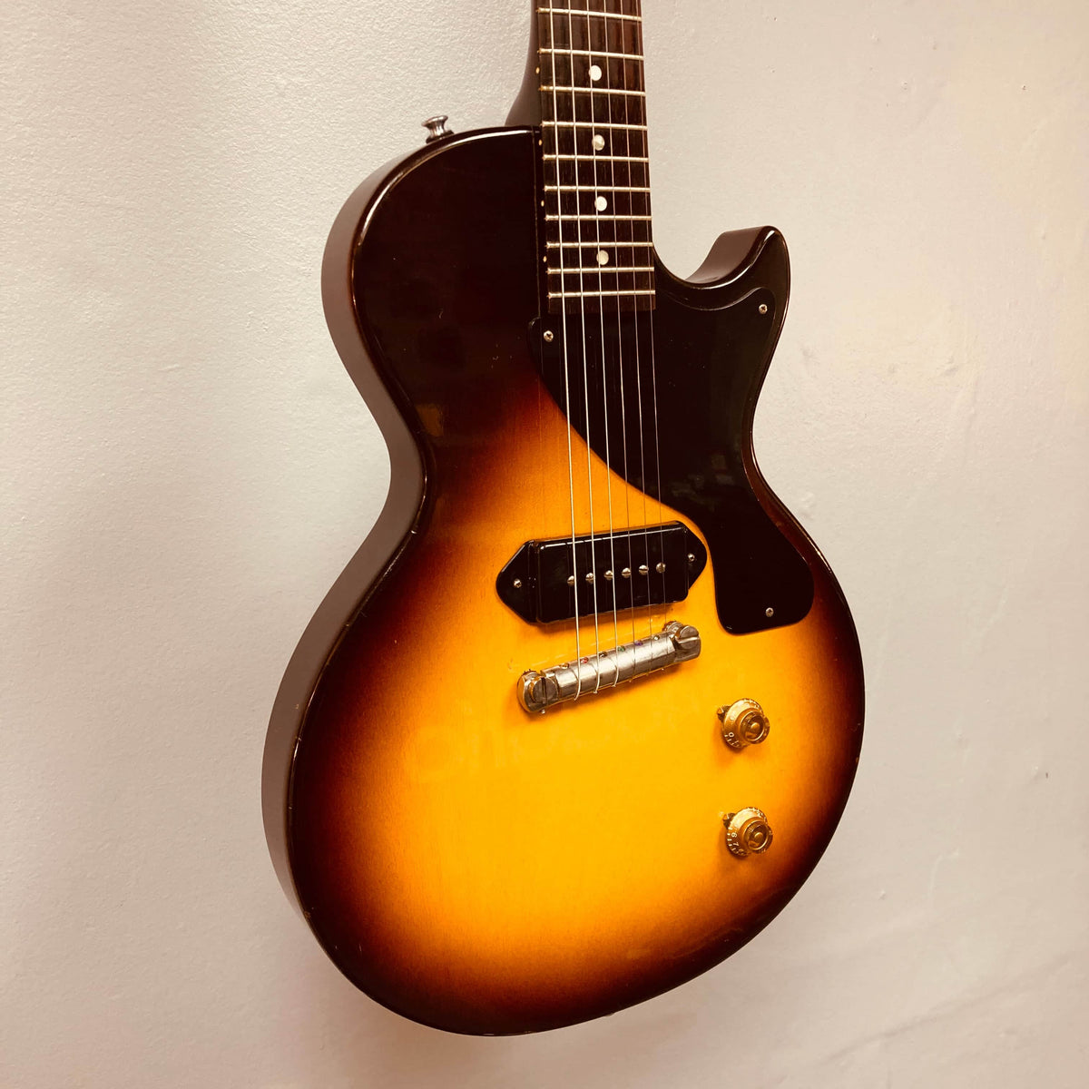 The Gibson Les Paul Junior 1957 Sunburst by Gibson features six strings, two control knobs, a single P-90 pickup, and a dark pickguard, displayed against a plain light-colored wall.