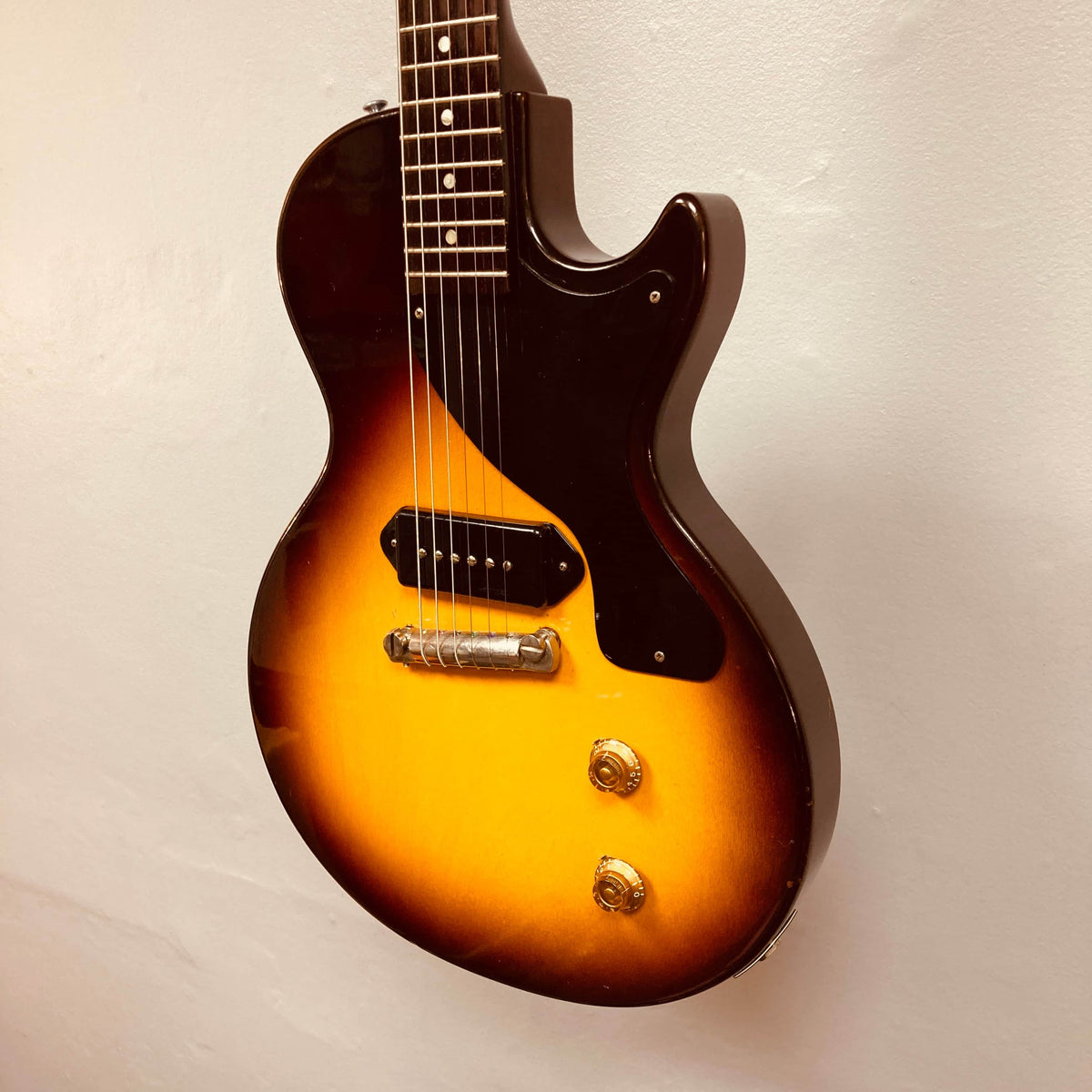 The Gibson Les Paul Junior 1957 Sunburst electric guitar features a single cutaway, six strings, two control knobs, and a black P-90 pickup, displayed against a plain light-colored wall.