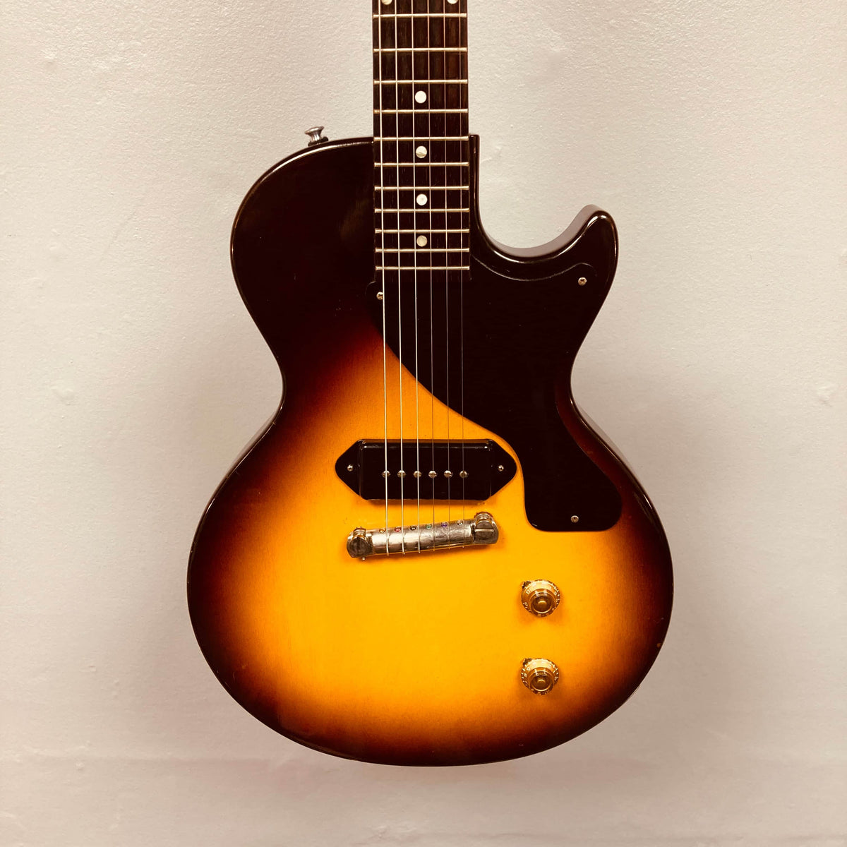 A Gibson Les Paul Junior 1957 Sunburst electric guitar with a single cutaway, two control knobs, a P-90 pickup, and a dark pickguard stands against a plain light wall.