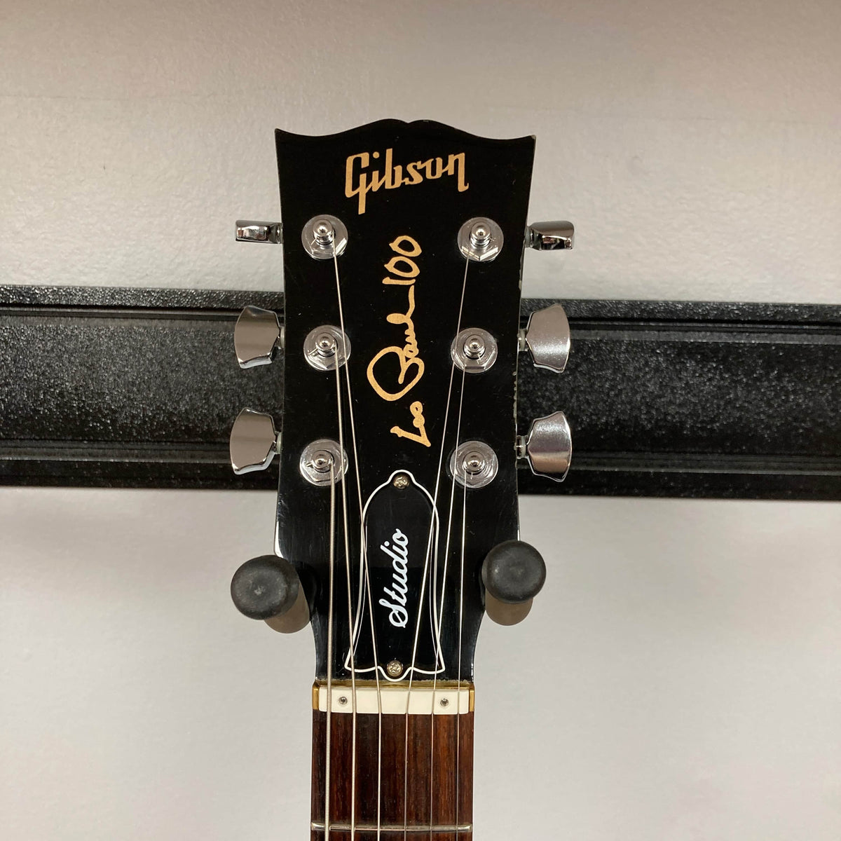 Close-up of a Gibson Les Paul Studio 2015 Desert Burst headstock, showing gold "Les Paul 100" script, six locking tuners, and strings, mounted on a wall hanger. Includes case. Brand: Gibson.