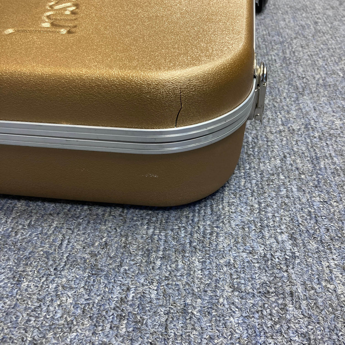 A brown hard-shell case with a cracked corner sits on a blue textured carpet; it once held a Gibson Les Paul Studio 2015 Desert Burst by Gibson, complete with upgraded locking tuners.