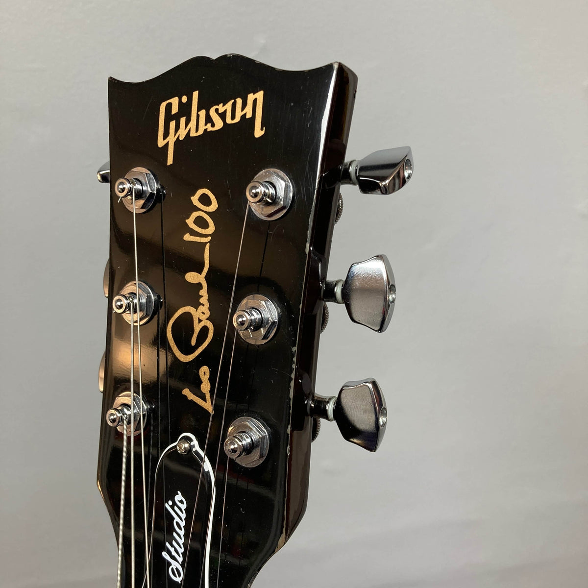 Close-up of a Gibson Les Paul Studio 2015 Desert Burst headstock with gold "Les Paul 100" signature, upgraded locking tuners, and an adjustable nut against a plain gray background. Brand: Gibson.