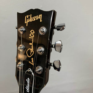Close-up of a Gibson Les Paul Studio 2015 Desert Burst headstock with gold "Les Paul 100" signature, upgraded locking tuners, and an adjustable nut against a plain gray background. Brand: Gibson.