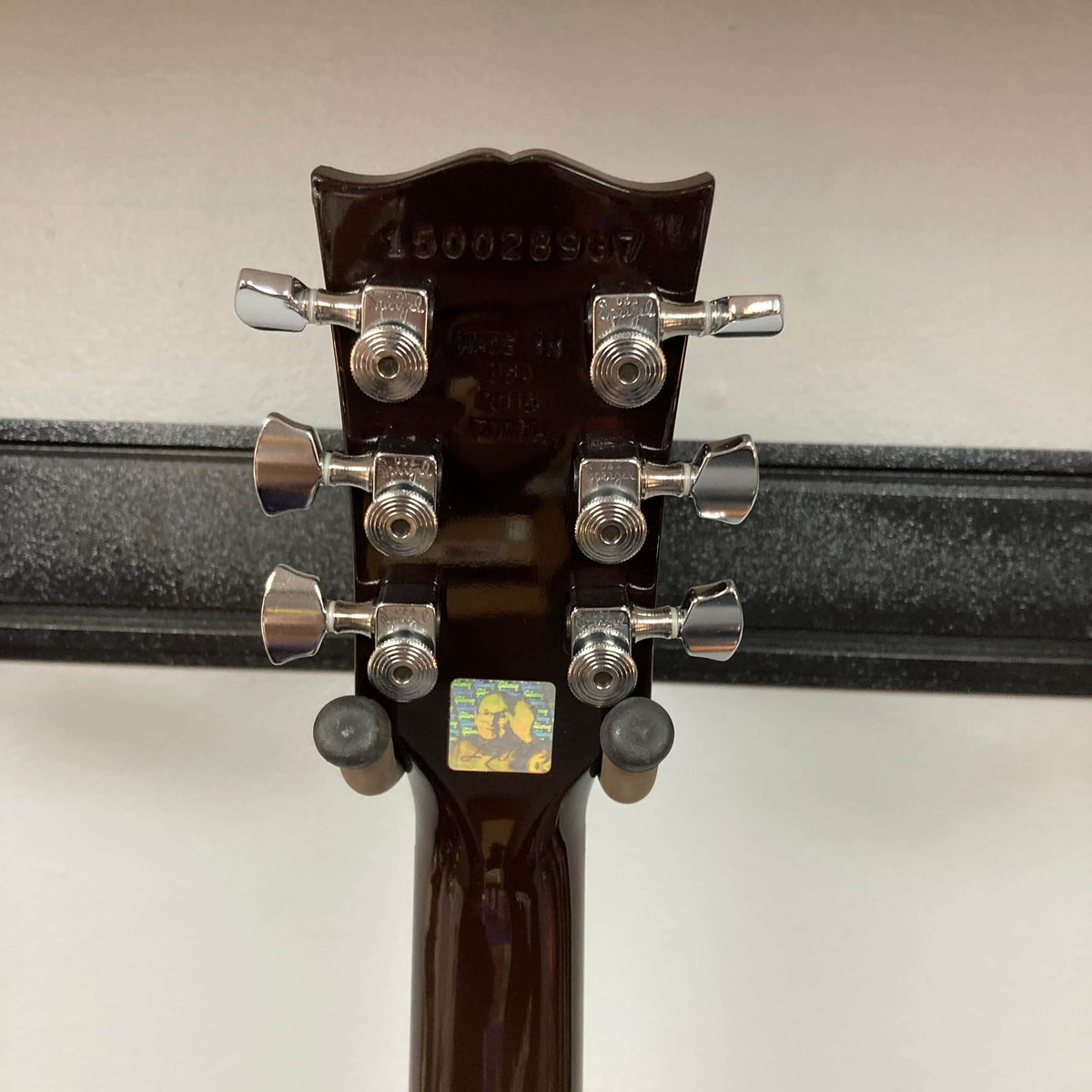 Close-up of the back of a Gibson Les Paul Studio 2015 Desert Burst headstock featuring six upgraded locking tuners, serial number, manufacturer info, and a small sticker with a graphic. Includes case. Brand: Gibson.