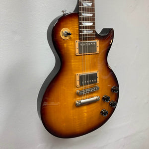 A Gibson Les Paul Studio 2015 Desert Burst electric guitar with dual humbuckers, locking tuners, four control knobs, and a three-way toggle is mounted on a light-colored wall; Gibson hard case included.