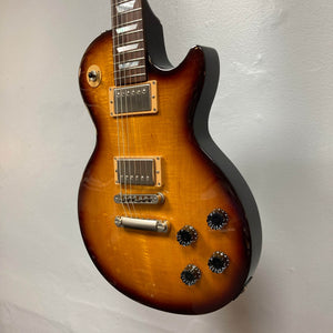 A Gibson Les Paul Studio 2015 Desert Burst electric guitar by Gibson, featuring two humbuckers, locking tuners, four black knobs, and a three-way switch, hangs on a plain light-colored wall.
