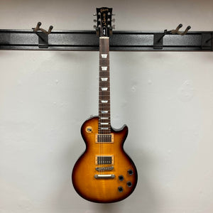 A Gibson Les Paul Studio 2015 Desert Burst electric guitar with dual humbuckers, four control knobs, upgraded locking tuners, and a case is hanging on a wall-mounted rack.