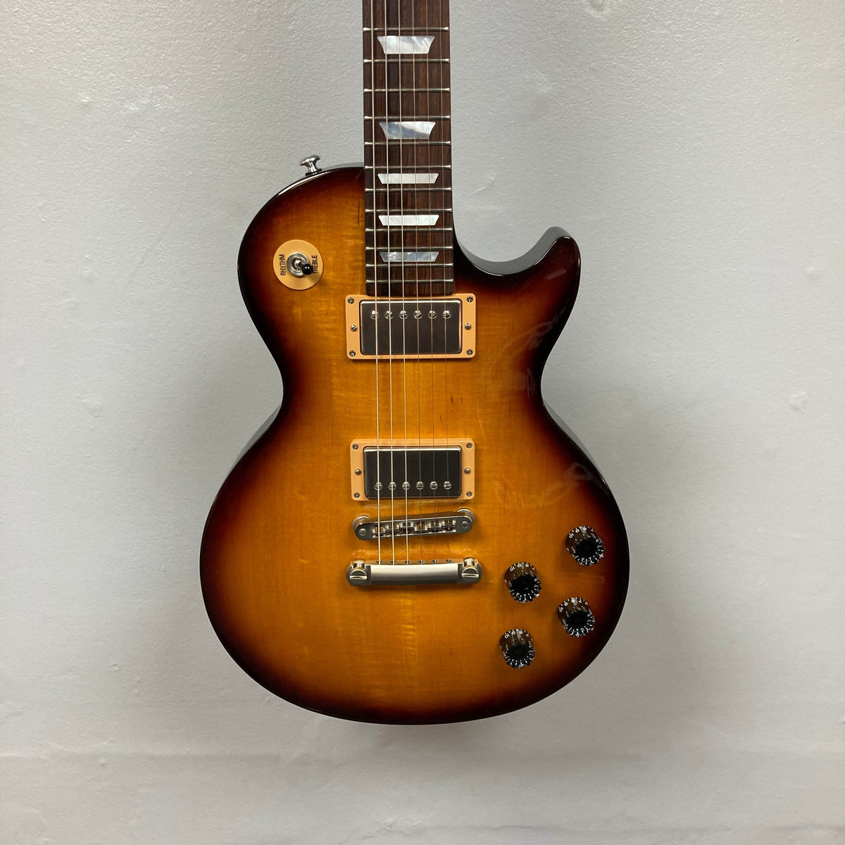 A Gibson Les Paul Studio 2015 Desert Burst electric guitar with two humbucker pickups, four knobs, locking tuners, and a single cutaway, shown against a plain light gray wall. Includes case. Brand: Gibson.