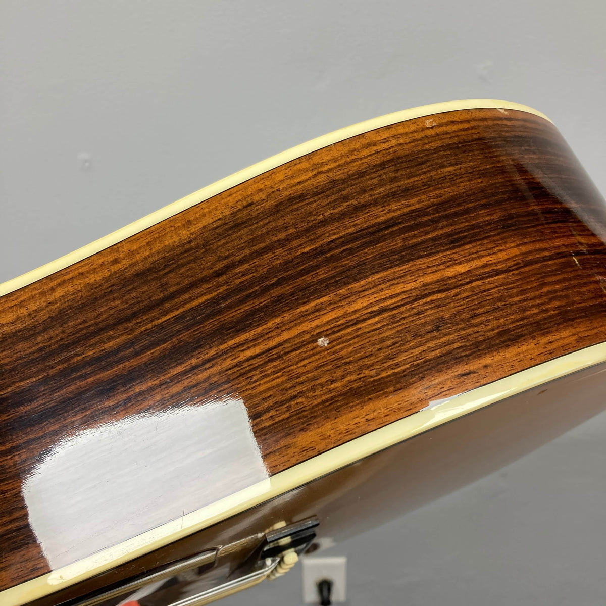 A close-up of a Gibson Nouveau NV6-R 1986 acoustic guitar’s side shows wood grain, cream binding, and a small dent, set against a gray wall.