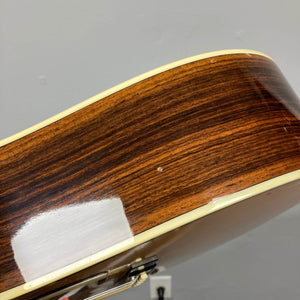 A close-up of a Gibson Nouveau NV6-R 1986 acoustic guitar’s side shows wood grain, cream binding, and a small dent, set against a gray wall.