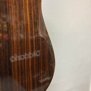 Close-up of the side of a Gibson Nouveau NV6-R 1986 acoustic guitar, highlighting its wood grain finish and square shoulder dreadnought body against a plain light-colored wall.