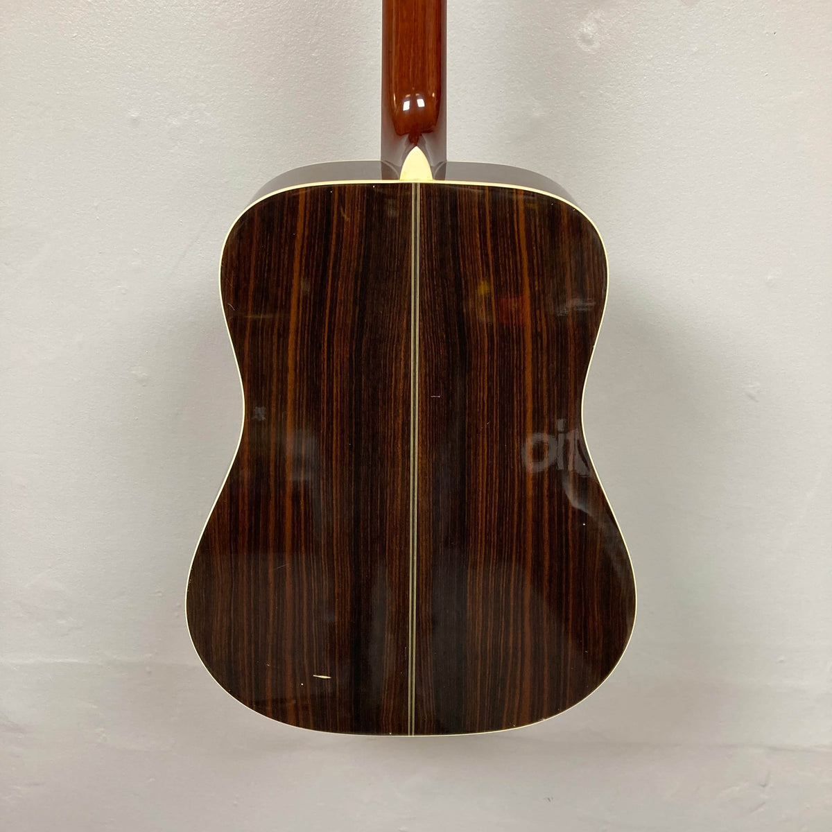 The back of a Gibson Nouveau NV6-R 1986 acoustic guitar, a square shoulder dreadnought by Gibson, features dark wood with a central lighter stripe and is displayed against a plain light-colored wall.