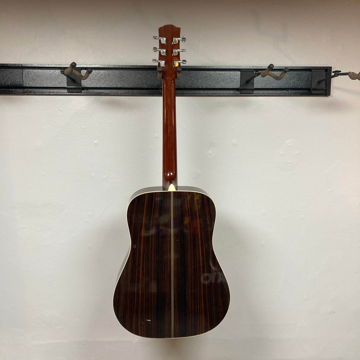 A Gibson Nouveau NV6-R 1986 acoustic guitar by Gibson hangs on a black wall-mounted rack with its back facing outward against a white wall, highlighting its classic square shoulder dreadnought design.