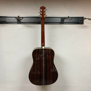 A Gibson Nouveau NV6-R 1986 acoustic guitar by Gibson hangs on a black wall-mounted rack with its back facing outward against a white wall, highlighting its classic square shoulder dreadnought design.