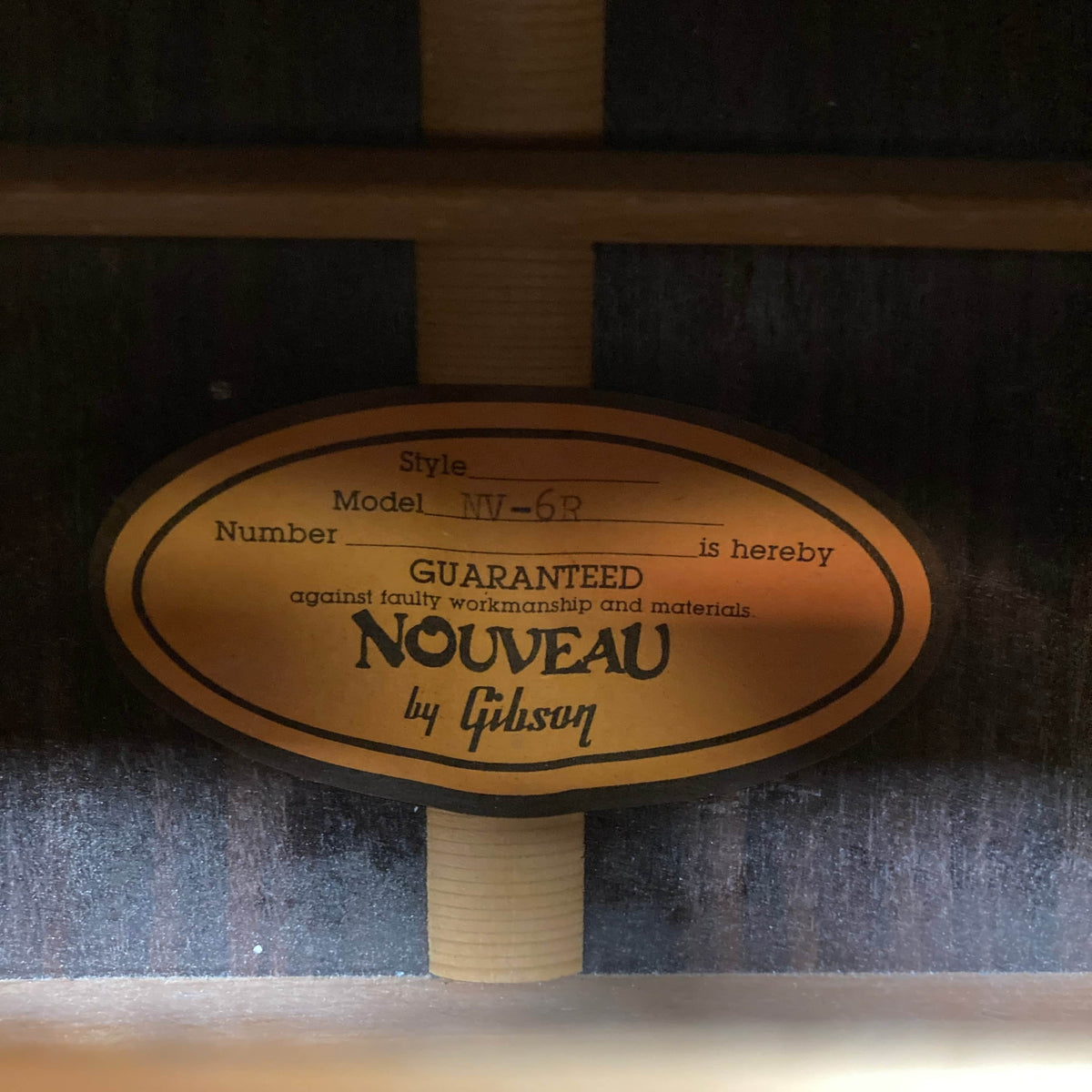 Close-up of the oval label inside a Gibson Nouveau NV6-R 1986 acoustic guitar, showing the model and stating it is "Guaranteed against faulty workmanship and materials" by Gibson.