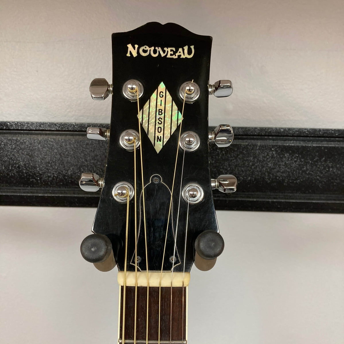 Close-up of a Gibson Nouveau NV6-R 1986 headstock, showing six tuning pegs and part of a guitar hanger against a light wall.