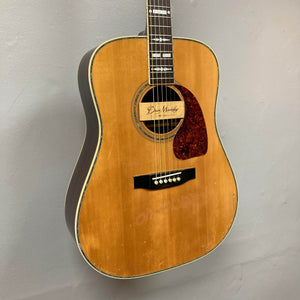 A Gibson Nouveau NV6-R 1986 acoustic square shoulder dreadnought with natural wood finish, tortoiseshell pickguard, and "Doc Murphy" signature on the soundhole label, reminiscent of a vintage guitar, hangs on a gray wall.