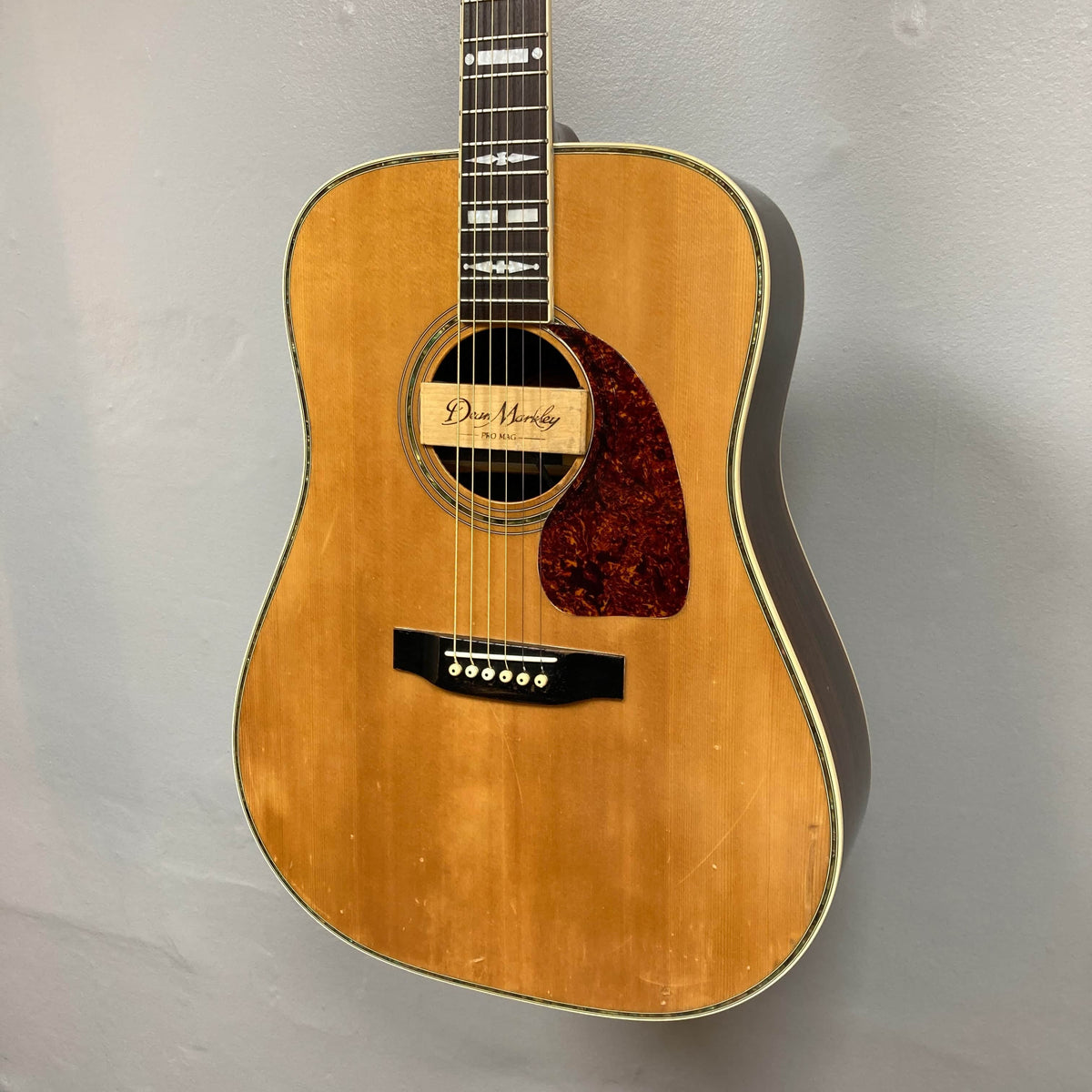 A Gibson Nouveau NV6-R 1986, a vintage square shoulder dreadnought with a tortoiseshell pickguard and signature inside the soundhole, hangs on a gray wall.