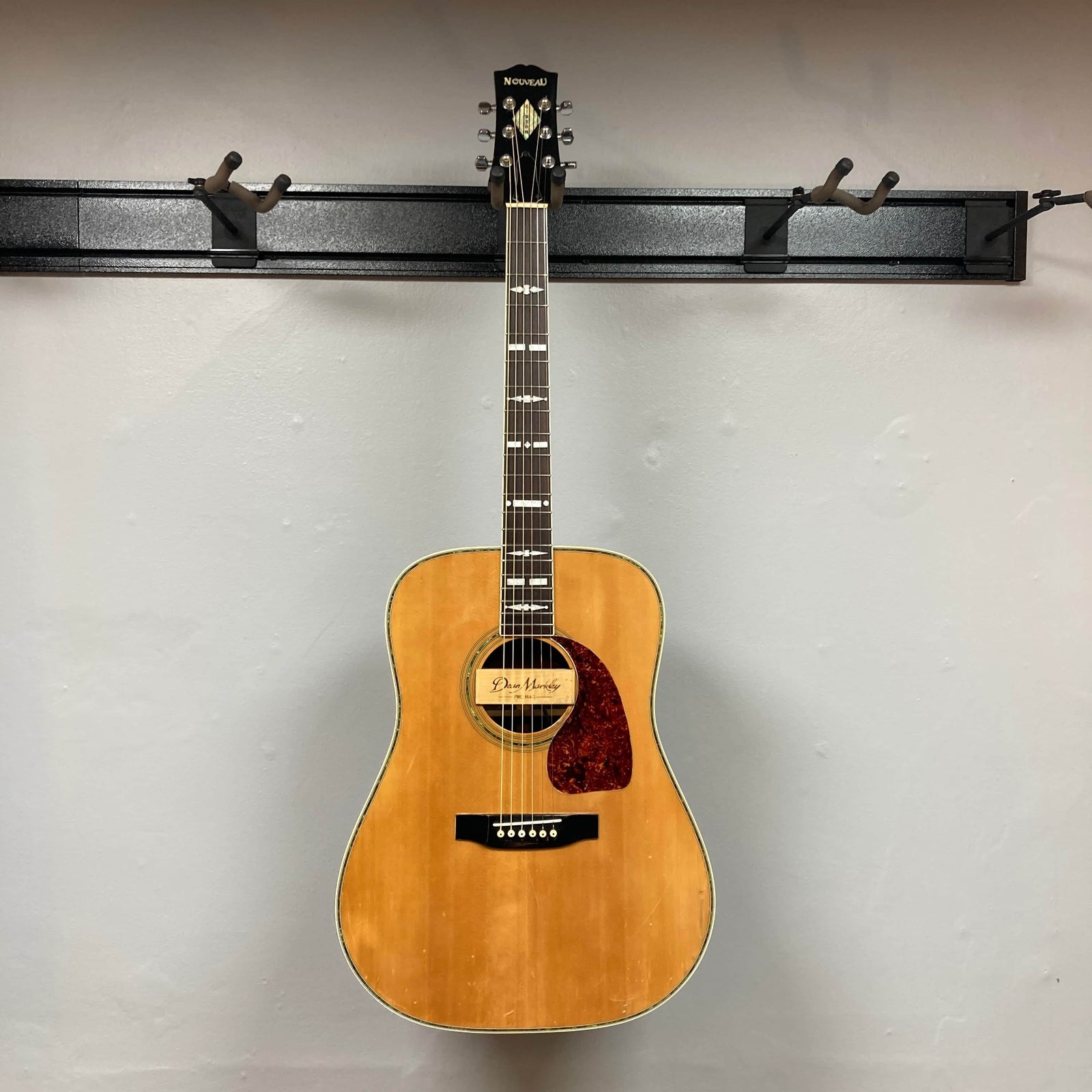 The Gibson Nouveau NV6-R 1986 is a vintage acoustic guitar with a natural wood finish, red pickguard, square shoulder dreadnought shape, and is pictured against a gray background.
