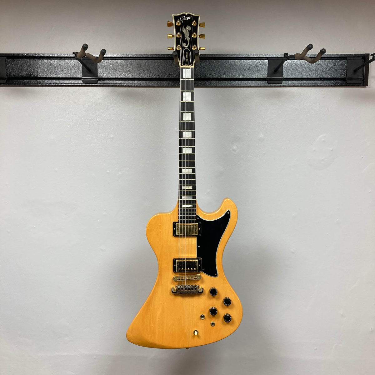 A Gibson RD Artist 1977–1982 vintage electric guitar with a natural finish, angular body, black pickguard, and six knobs hangs on a wall rack. Available from Guitars on Main with case included.
