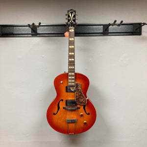 A Godin 5th Avenue Jumbo HB Memphis Sun hollow-body electric guitar hangs on a black wall-mounted rack.