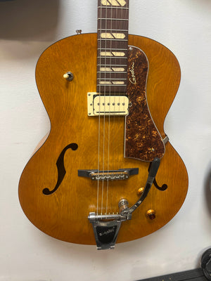 A Godin 5th Avenue Jumbo P-Rail Harvest Gold archtop guitar with a natural finish, single pickup, tortoiseshell pickguard, and Bigsby-style tailpiece hangs on a white wall.