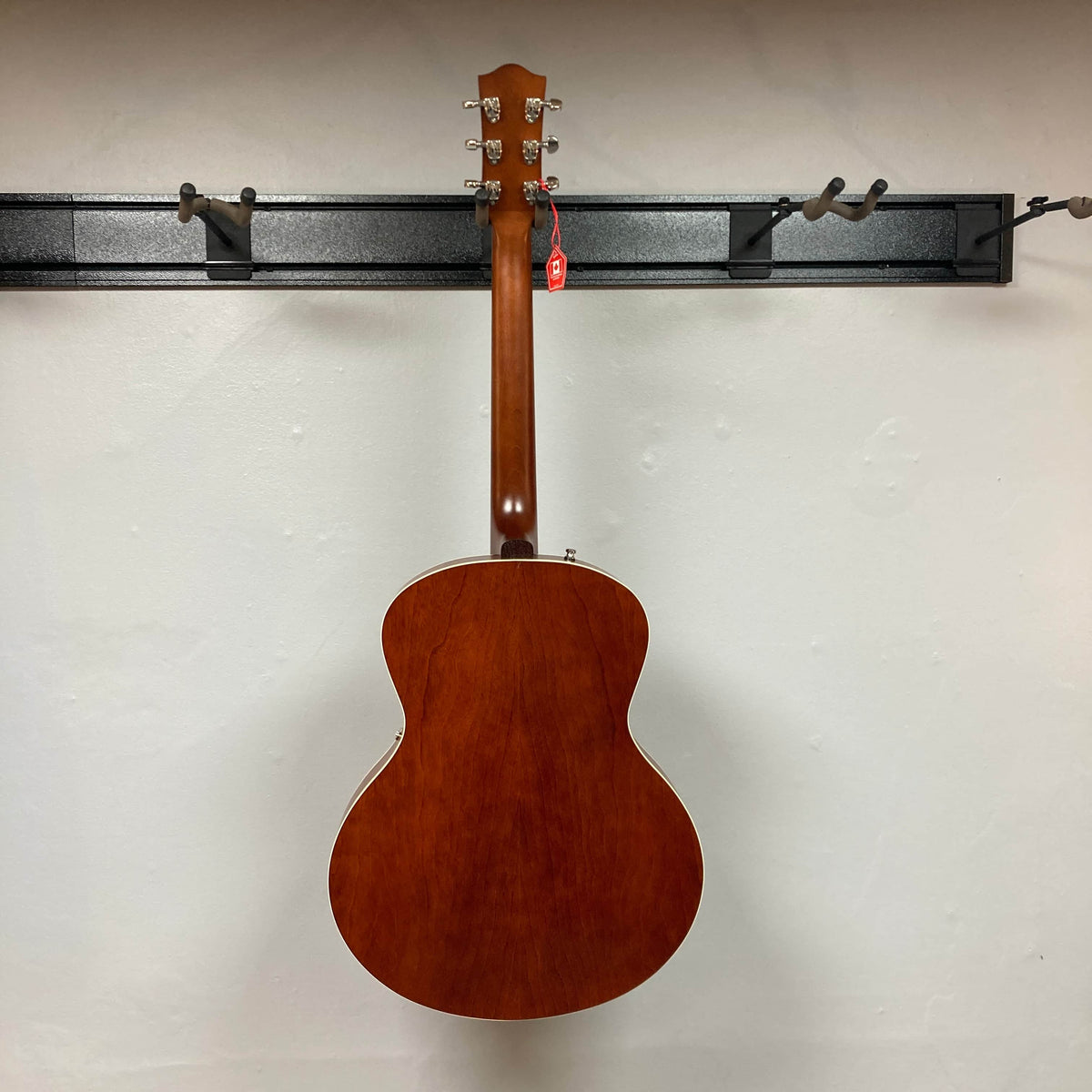 A Godin 5th Avenue Kingpin P90 Cognac Burst SF hangs on a wall rack, its wooden finish and red tag standing out against a light wall to showcase the vintage appeal of this renowned Godin acoustic guitar.