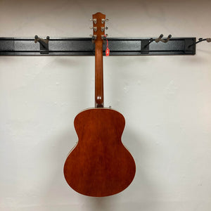 A Godin 5th Avenue Kingpin P90 Cognac Burst SF hangs on a wall rack, its wooden finish and red tag standing out against a light wall to showcase the vintage appeal of this renowned Godin acoustic guitar.