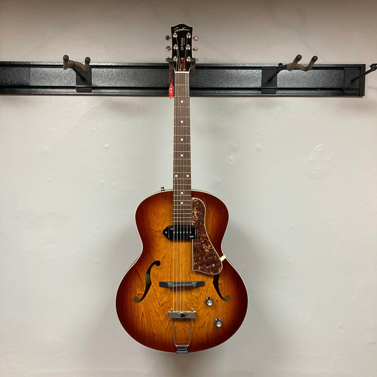 A Godin 5th Avenue Kingpin P90 Cognac Burst SF archtop electric guitar with dual control knobs and f-hole design hangs on a wall-mounted black rack, showcasing its vintage style and tone.
