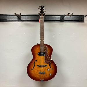A Godin 5th Avenue Kingpin P90 Cognac Burst SF archtop electric guitar with dual control knobs and f-hole design hangs on a wall-mounted black rack, showcasing its vintage style and tone.
