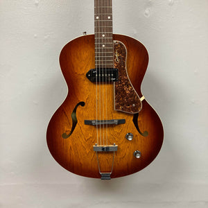 A Godin 5th Avenue Kingpin P90 Cognac Burst SF archtop electric guitar with dual f-holes, two knobs, a wooden pickguard, and visible wood grain hangs against a light wall, ready to deliver classic vintage tones.