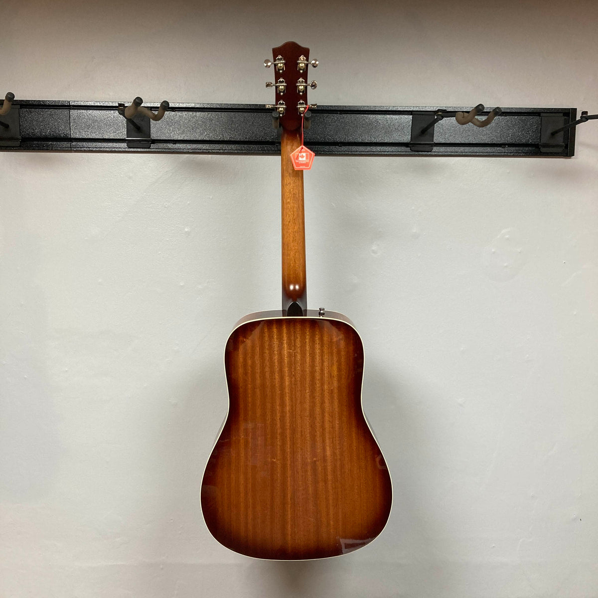 A Godin Century Maho Rustic Burst EQ SF acoustic guitar in brown, seen from the back, hangs on a black wall-mounted rack against a plain white wall.