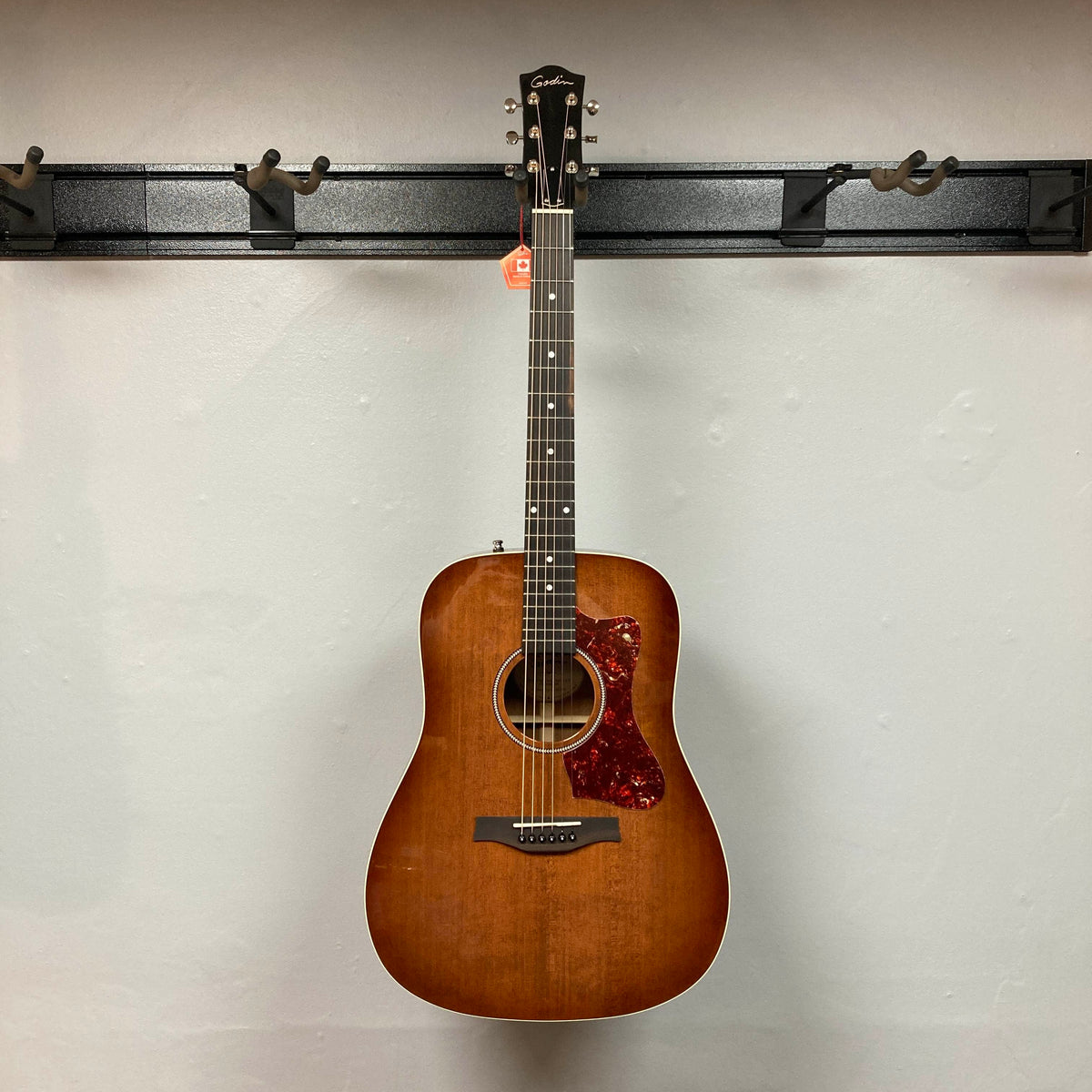 A Godin Century Maho Rustic Burst EQ SF by Godin, a dreadnought acoustic-electric guitar with a brown sunburst finish, hangs on a wall-mounted rack. The guitar features six strings, a pickguard, and is crafted from solid wood—blending classic aesthetics with modern electronics for versatile performance.