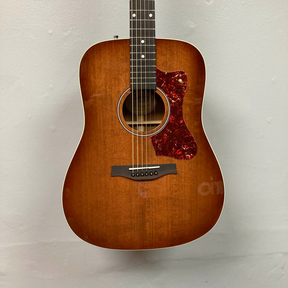 A Godin Century Maho Rustic Burst EQ SF solid wood guitar with a red tortoiseshell pickguard hangs on a plain light-colored wall.