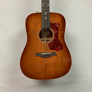 A Godin Century Maho Rustic Burst EQ SF solid wood guitar with a red tortoiseshell pickguard hangs on a plain light-colored wall.