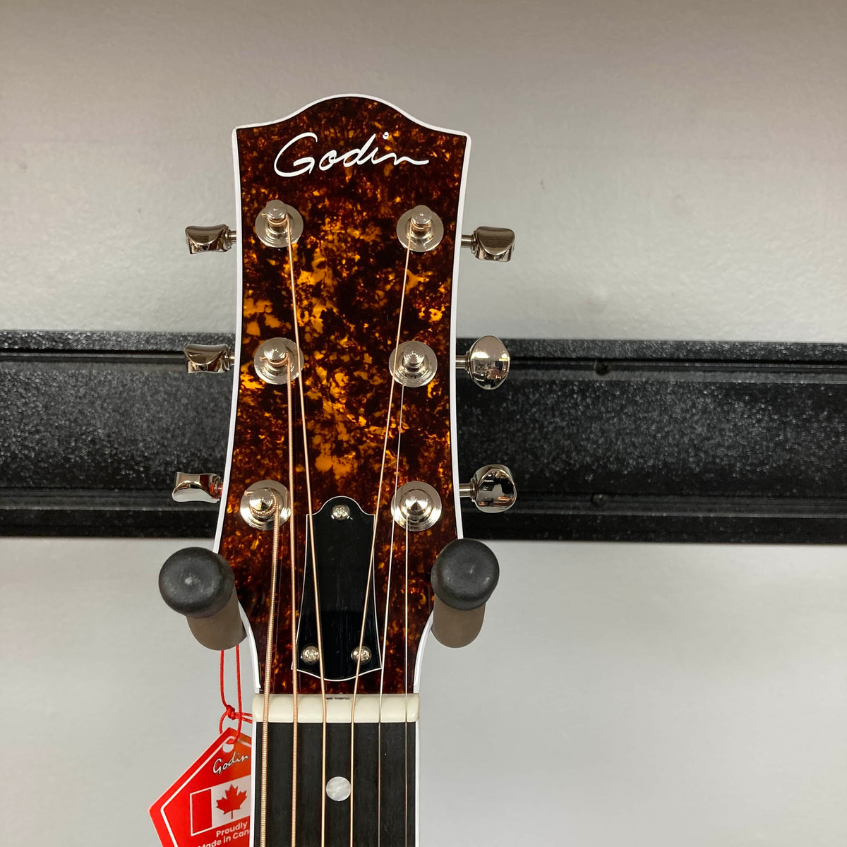 Close-up of the Godin Imperial Laguna Blue GT EQ SF Acoustic Guitar headstock with tuning pegs and strings, set against a gray wall. A red Canadian flag tag hangs from the neck. Includes gigbag.