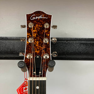 Close-up of the Godin Imperial Laguna Blue GT EQ SF Acoustic Guitar headstock with tuning pegs and strings, set against a gray wall. A red Canadian flag tag hangs from the neck. Includes gigbag.