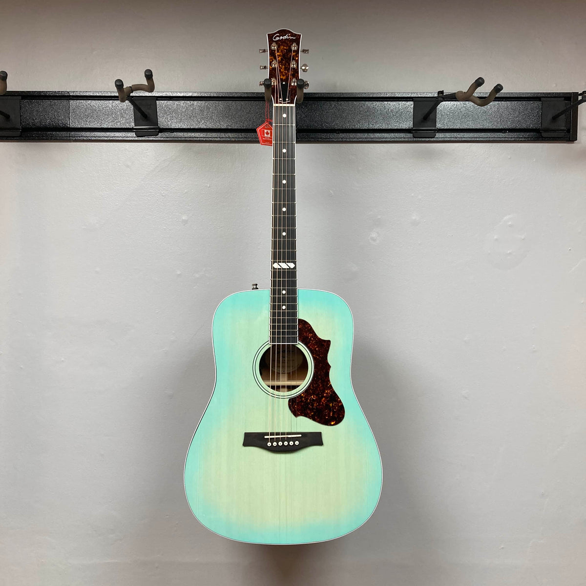 A Godin Imperial Laguna Blue GT EQ SF Acoustic Guitar w/Gigbag hangs on a wall-mounted holder against a plain white wall.