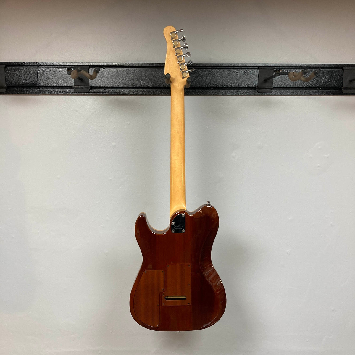 A Godin Passion Custom Swamp Ash Electric Guitar w/ GigBag Consign is hanging on a wall-mounted rack, displaying its back and neck against a plain white wall.